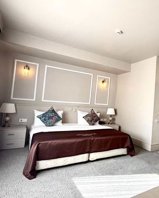 A hotel room featuring a bed with patterned carpet, brown textured walls, and a wooden luggage rack in the foreground.