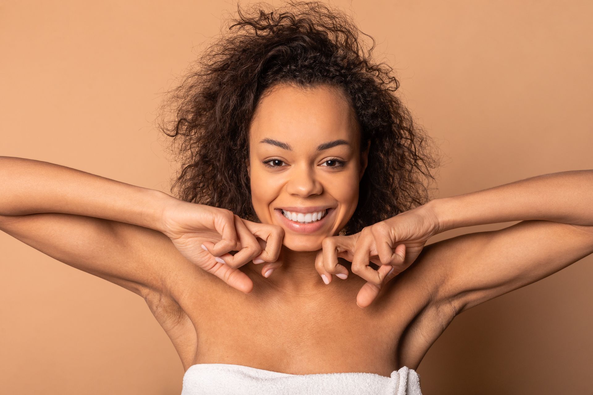 Smiling person with curly hair posing against a beige background, wearing a white towel top.