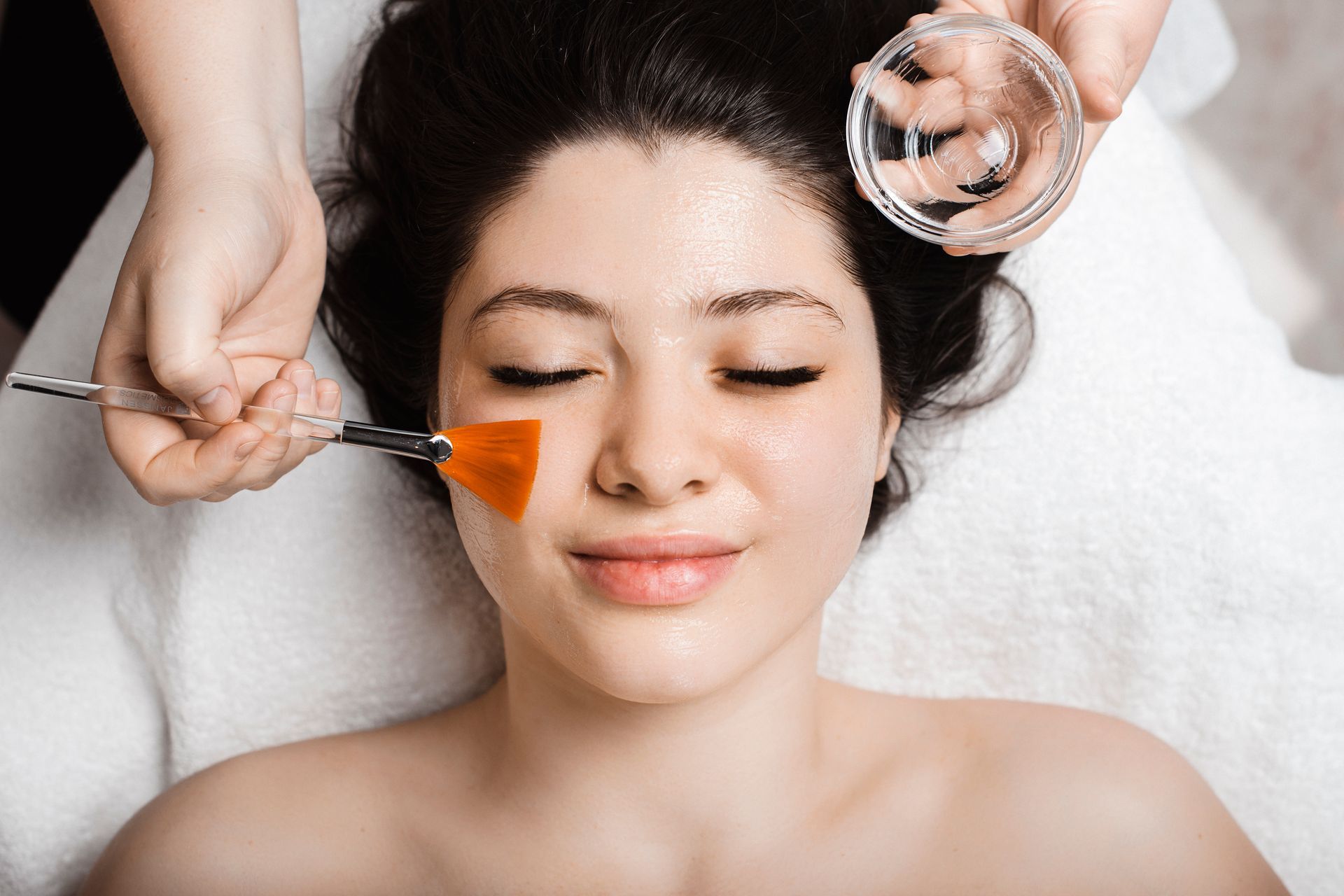 Facial treatment. A person reclines while a skincare professional applies a product with a brush and bowl.