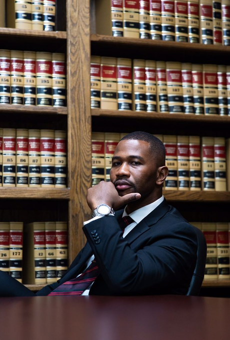 A man in a suit and tie is sitting at a desk in front of a bookshelf.