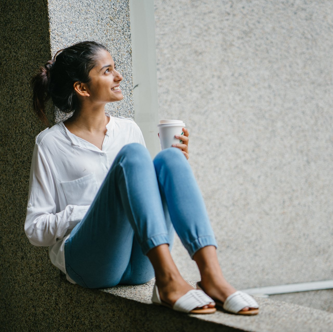 Eine Frau sitzt glücklich auf einem Fensterbrett und hält eine Tasse Kaffee
