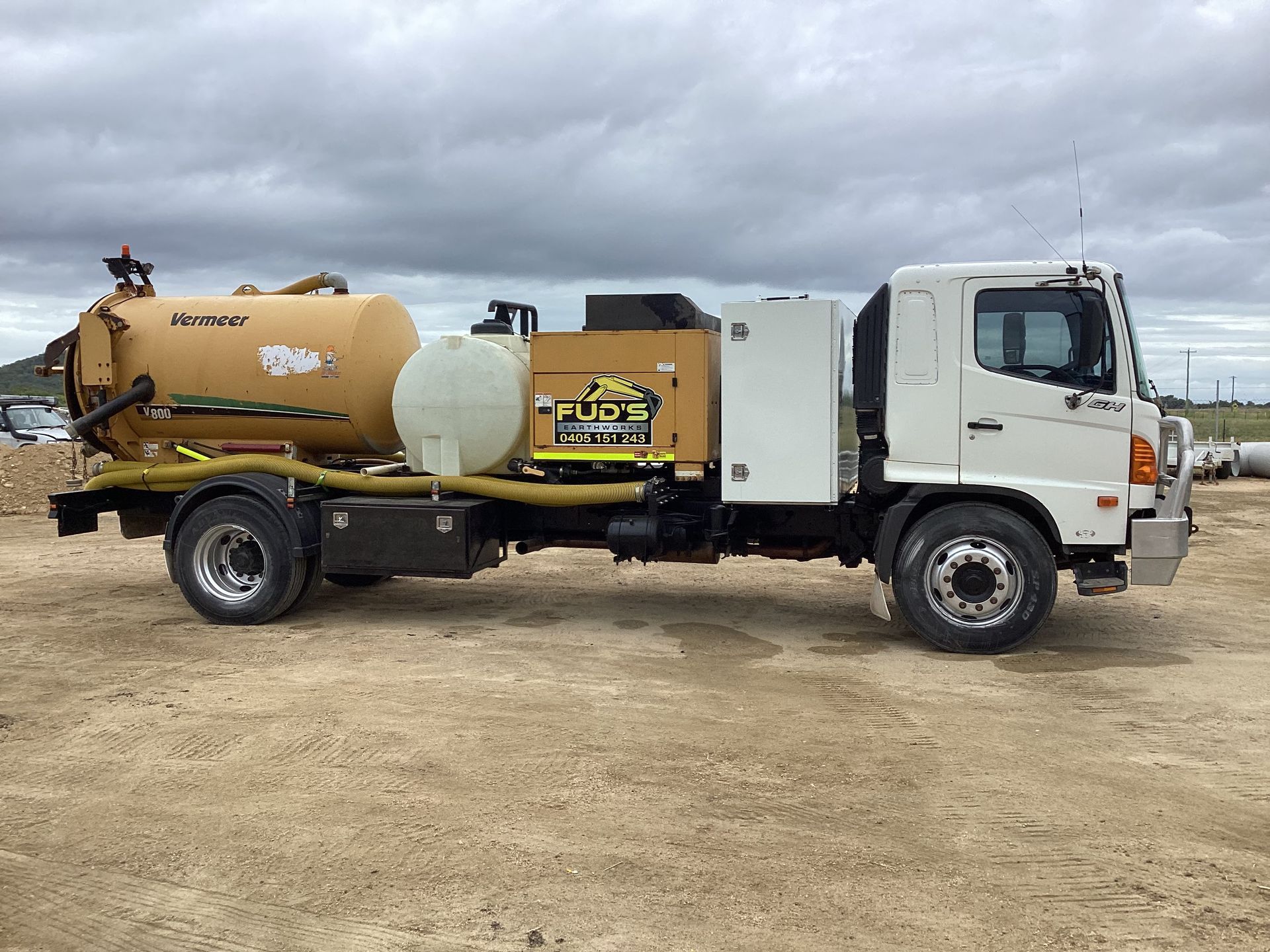 Yellow and white vacuum truck parked on a dirt lot under a cloudy sky