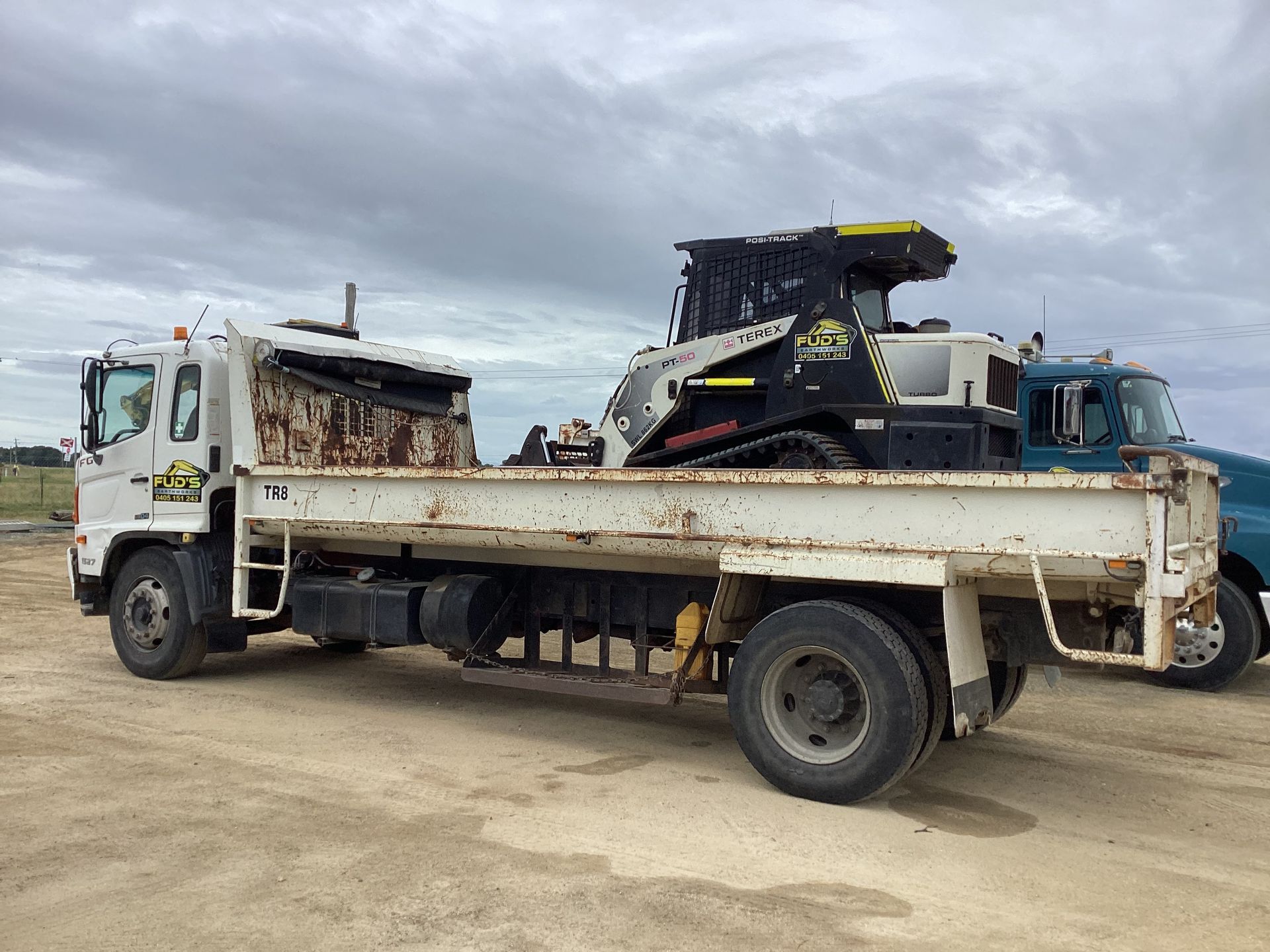 Rusty white flatbed truck hauling a large black and yellow machine on a dirt lot under cloudy skies