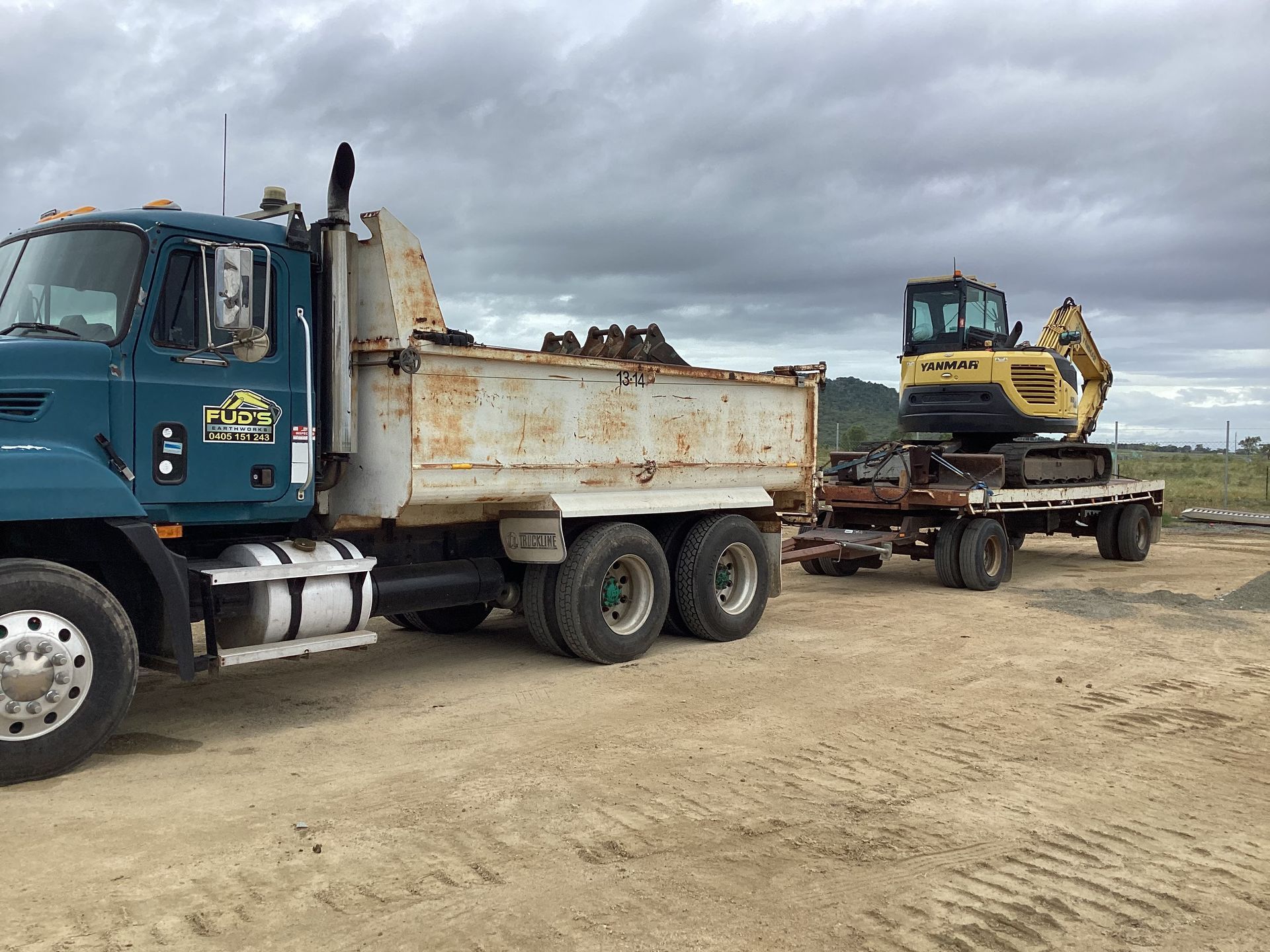 Blue dump truck hauling a yellow excavator on a flatbed in a muddy construction yard under cloudy skies