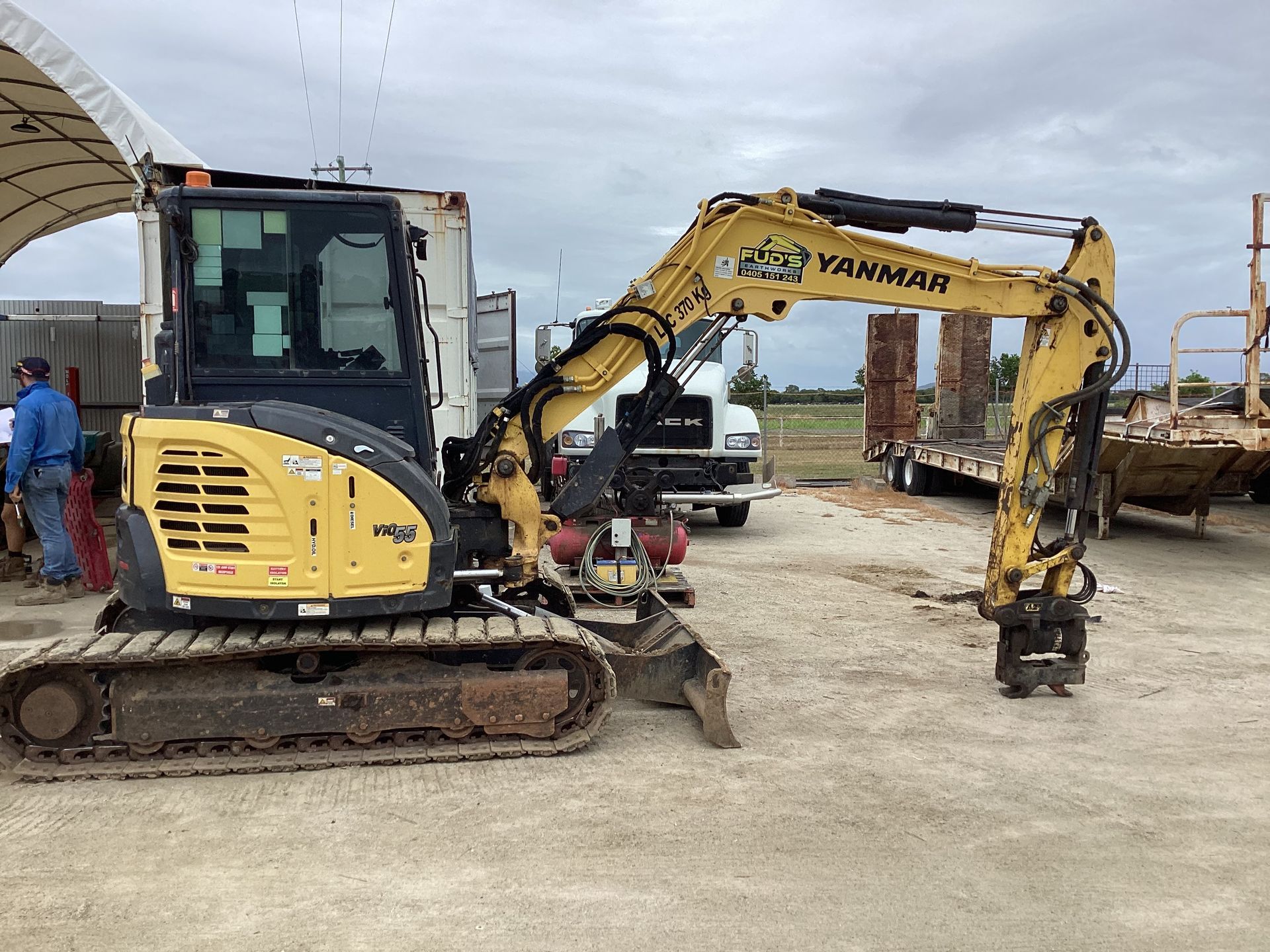 Yellow Yanmar mini excavator on a dirt lot, with another excavator and workers in the background.