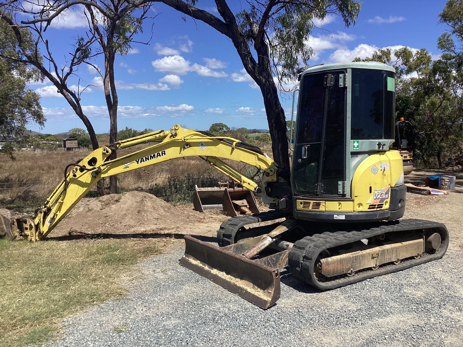 Yellow excavator parked by a gravel pile in a grassy field under trees and blue sky.