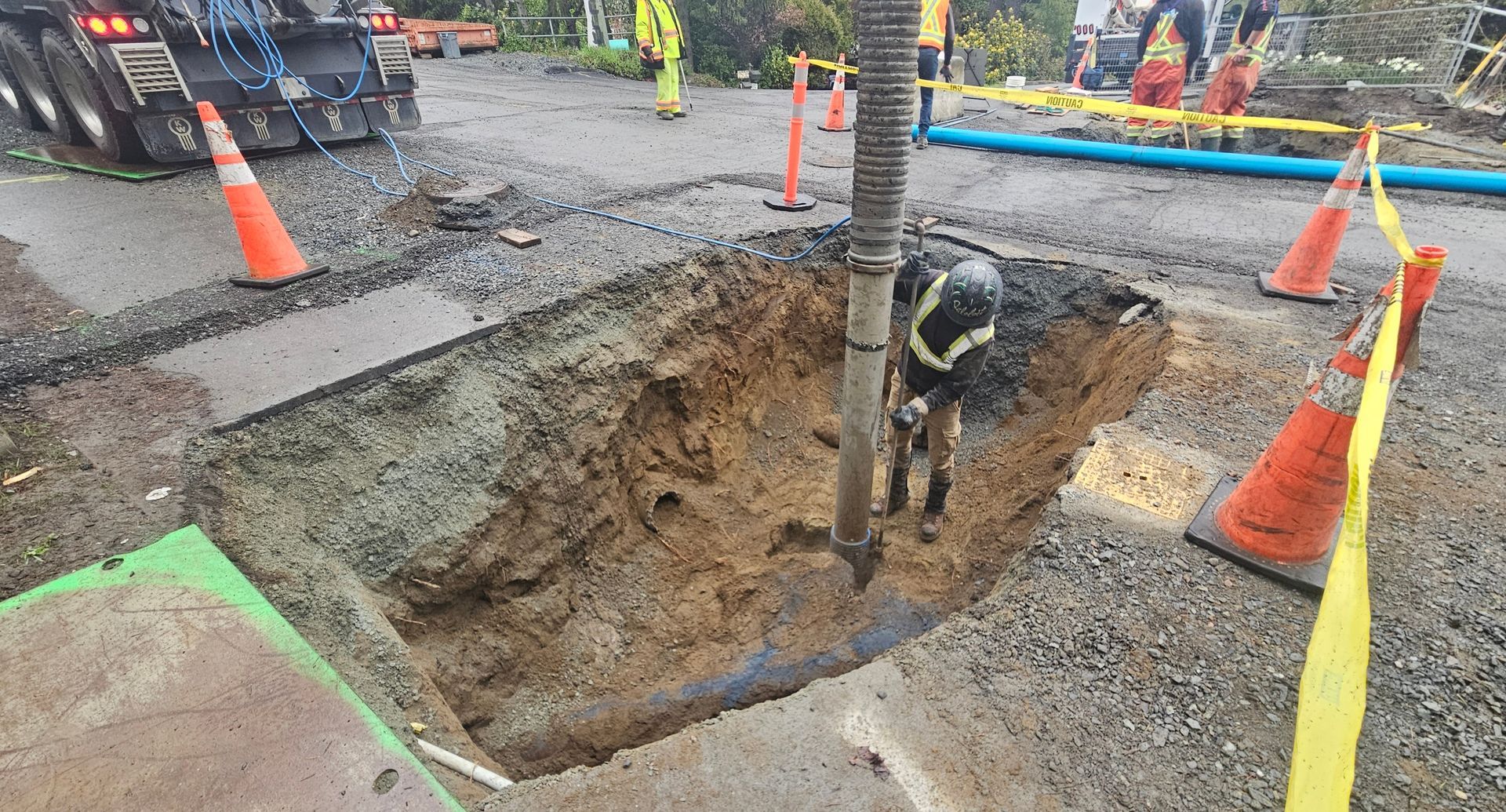 Roadwork excavation with a deep open trench, cones, caution tape, and a worker beside a utility pole.