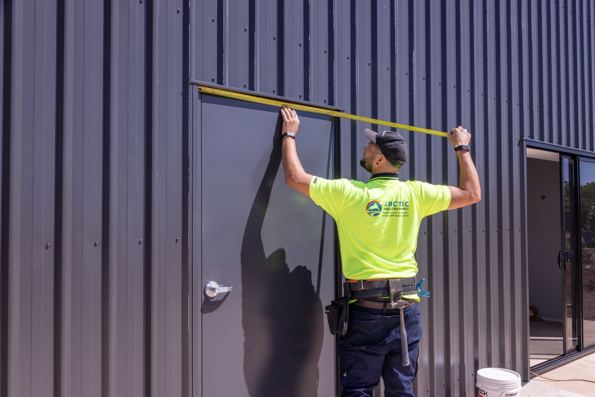Worker in a neon safety shirt measures a  corrugated metal wall with a tape measure. — Arctic Installations in Winnellie, NT