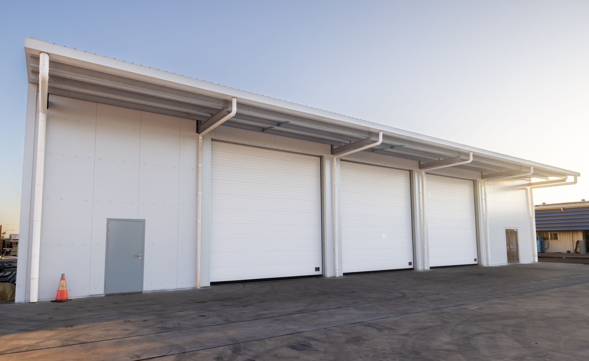 Industrial warehouse with white roll-up doors and a gray side door under a metal awning at dusk — Arctic Installations in Winnellie, NT