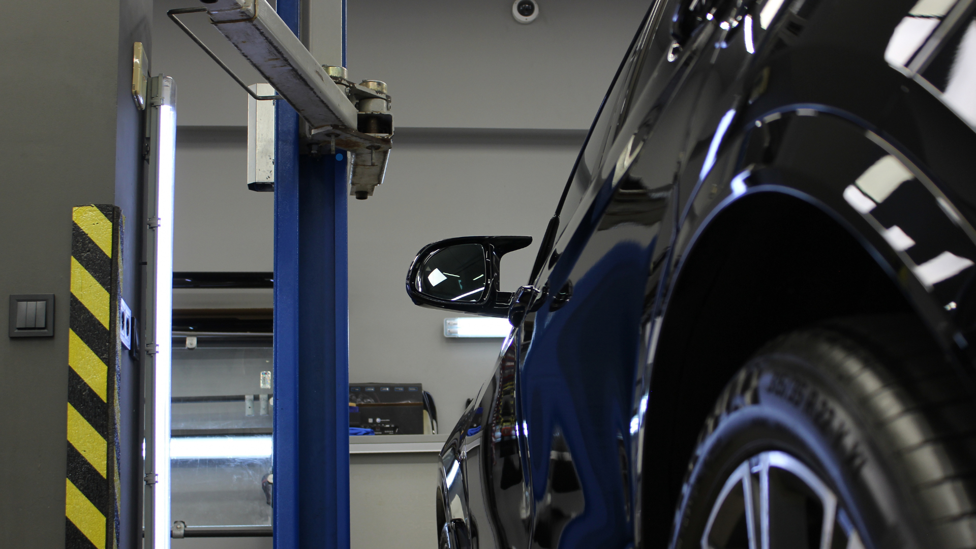 A black car's side view with the car on a lift, showing the sleek design of the car's exterior. The workshop is well-lit with a clean, modern setup, and the yellow and black safety stripes are visible on the wall.