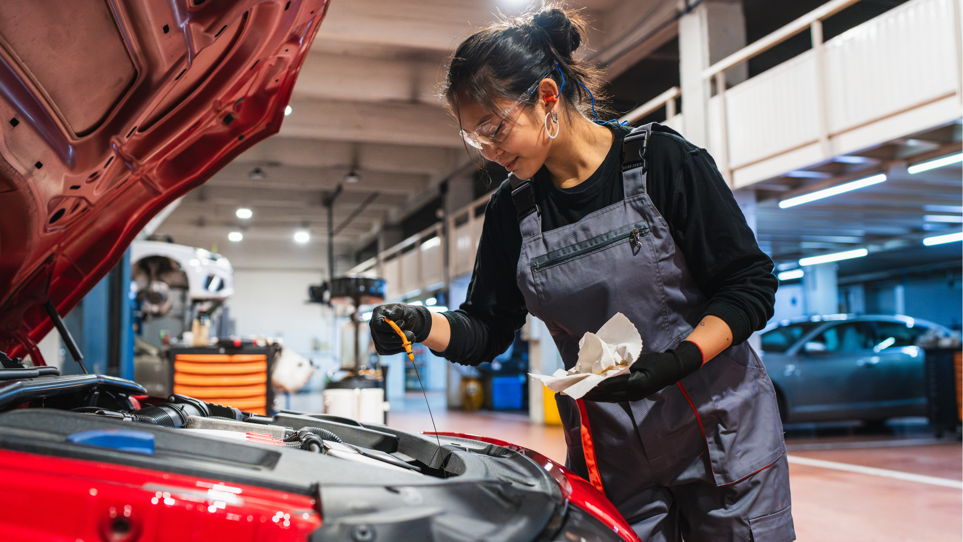 A female mechanic wearing safety glasses and a gray work overall checking the engine oil of a red car in a well-lit auto repair shop, highlighting professional car maintenance.
