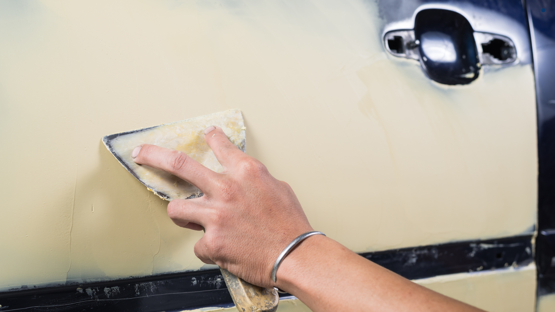 Technician hand sanding body filler on a car door panel during repair, smoothing surface imperfectio