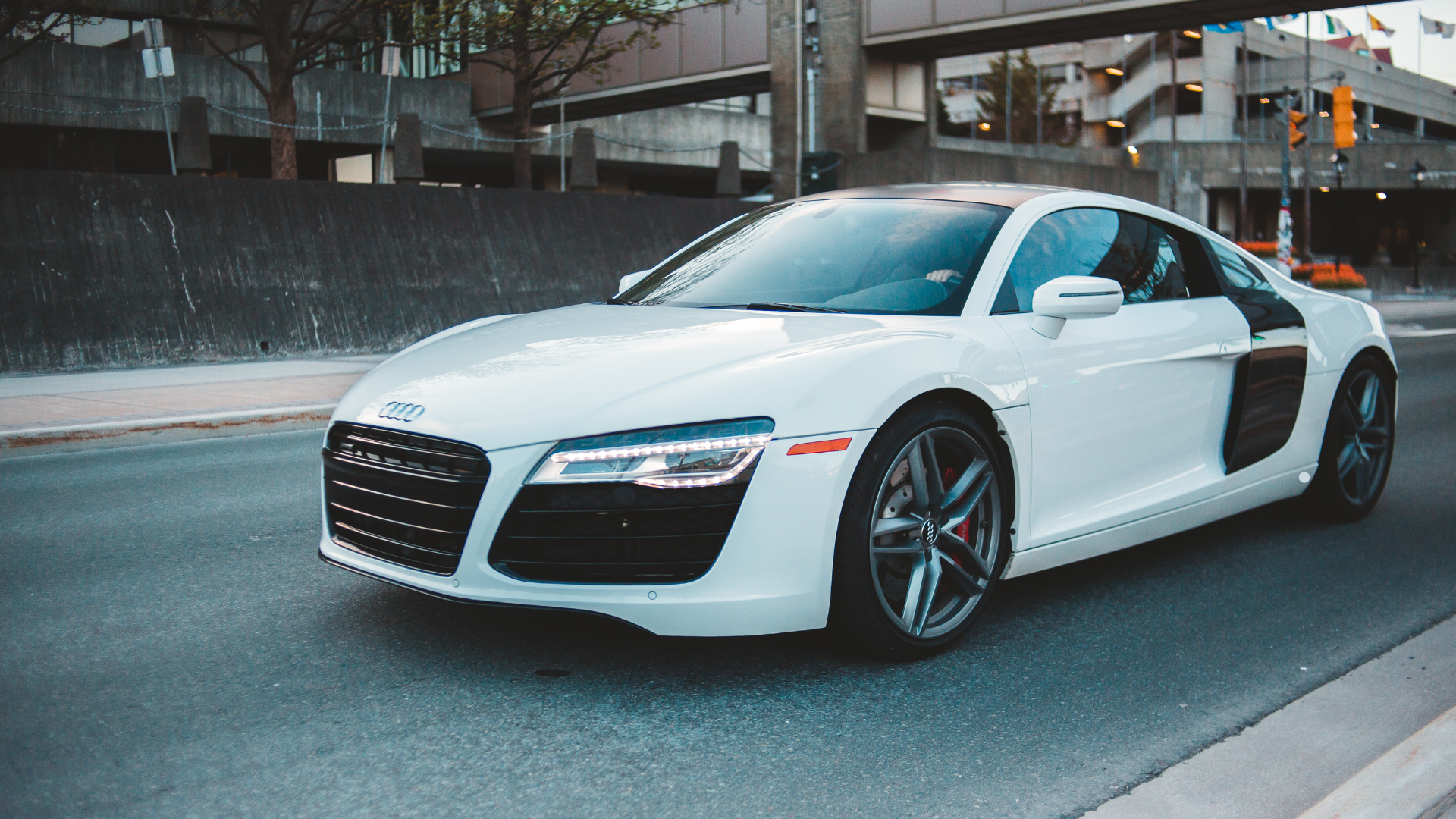 White high-end luxury sports car parked on a city street, showing the front and side profile with modern buildings in the background.