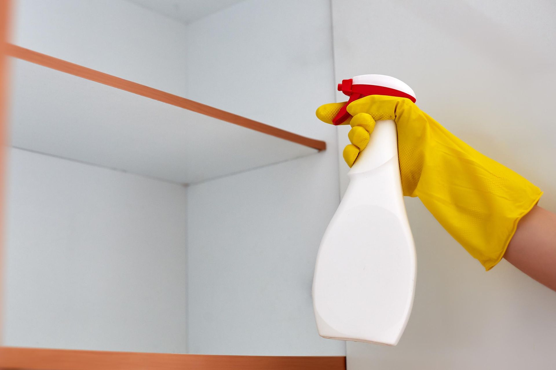 A person wearing yellow gloves is spraying a shelf with a spray bottle.