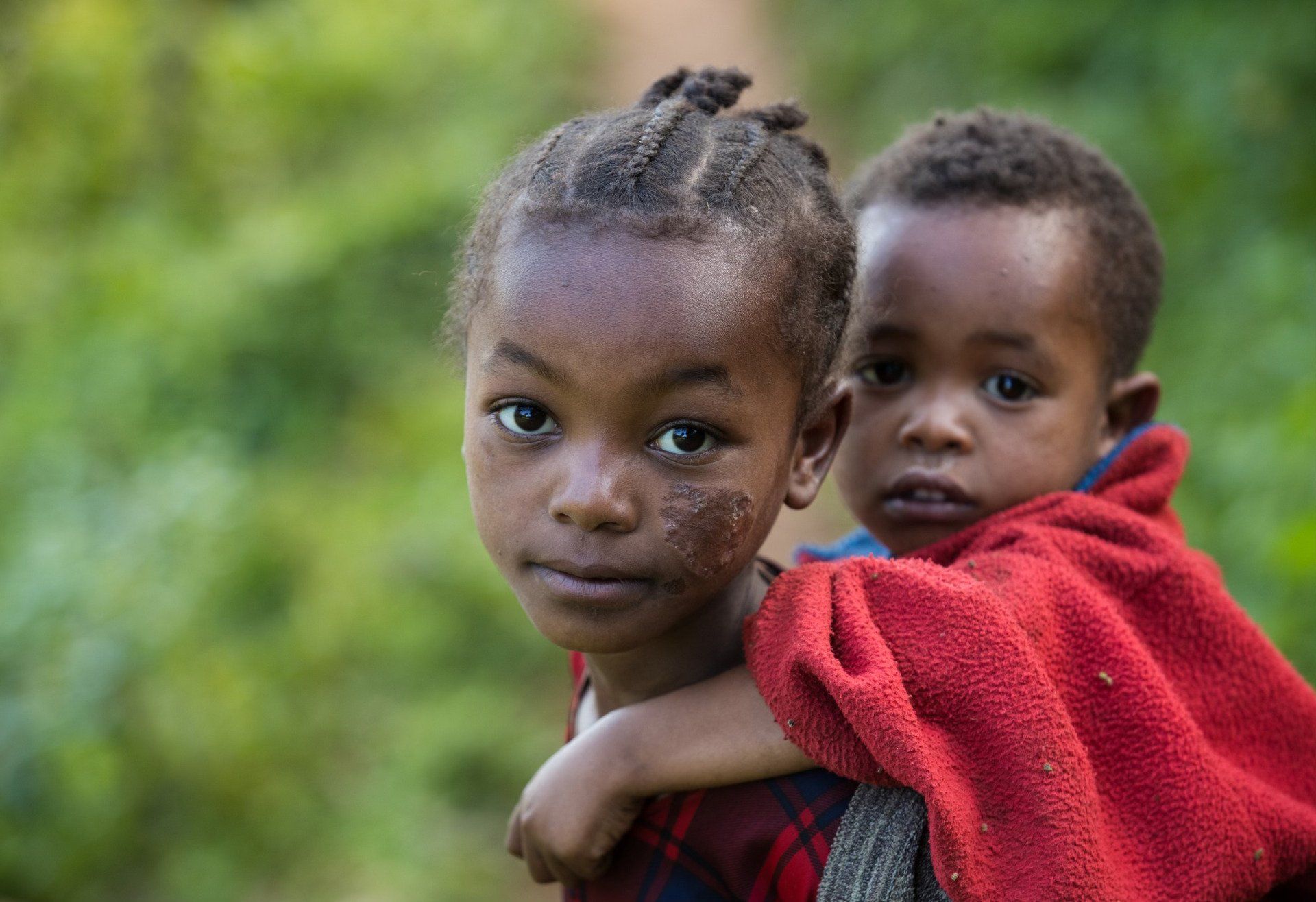 Two children in red clothing, one carrying the other, outdoors against a blurred green background