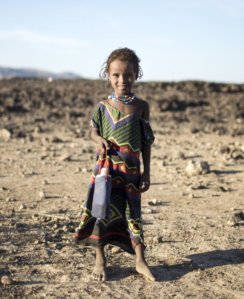 A child standing barefoot on rocky ground in a patterned dress, outdoors under a bright sky.