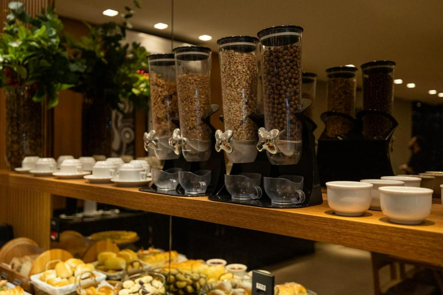 Buffet de café da manhã com dispensadores de cereais, xícaras e produtos de panificação em um balcão de madeira.