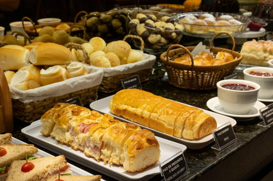 Buffet com uma variedade de pães, doces e acompanhamentos como geleias, em um ambiente com pouca luz.