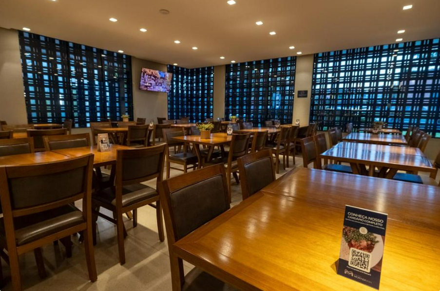 Sala de jantar com mesas e cadeiras de madeira, parede com destaque em tom escuro e uma TV.