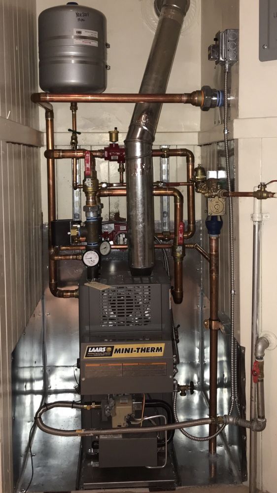 Residential boiler and pipe system in a utility room, with gray tank, black chimney flue, and copper pipes.