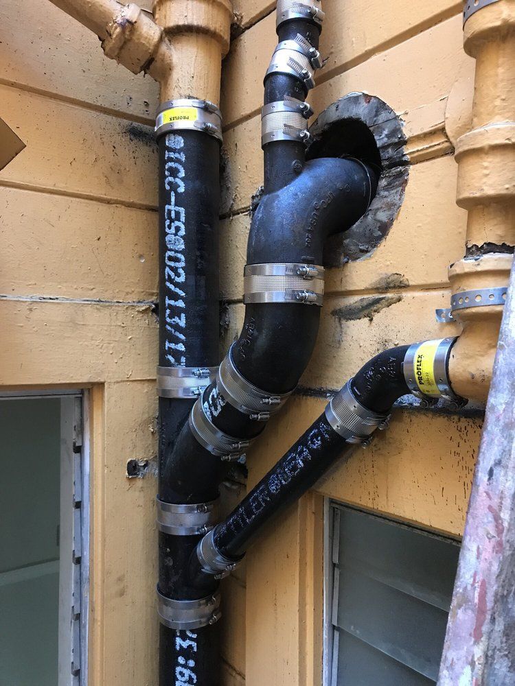 Black insulated pipes and ducts mounted on a tan exterior wall with metal clamps and a window below