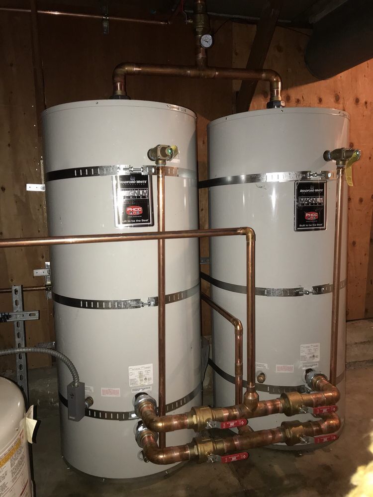 Two white water heater tanks in a utility closet with copper pipes and valves.