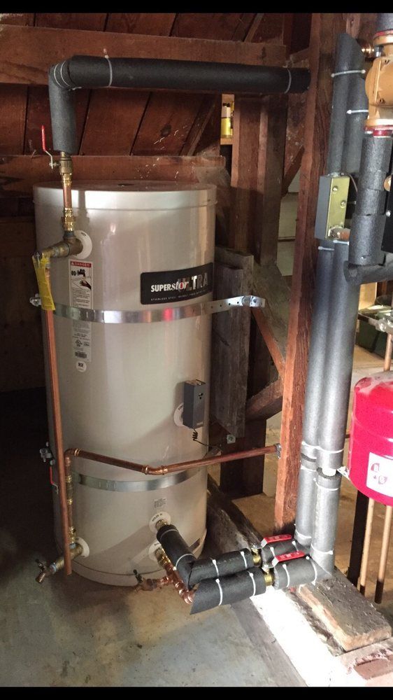 Vertical silver water heater with black insulated pipes in a basement utility area