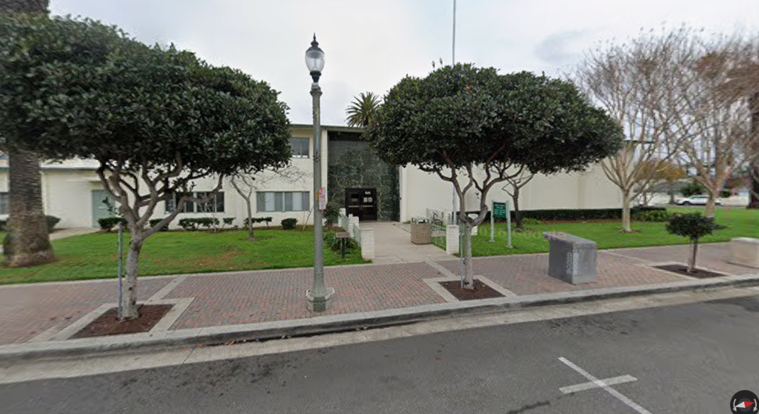 Street view of a building entrance with trimmed trees, a lamppost, and sidewalk along the curb.