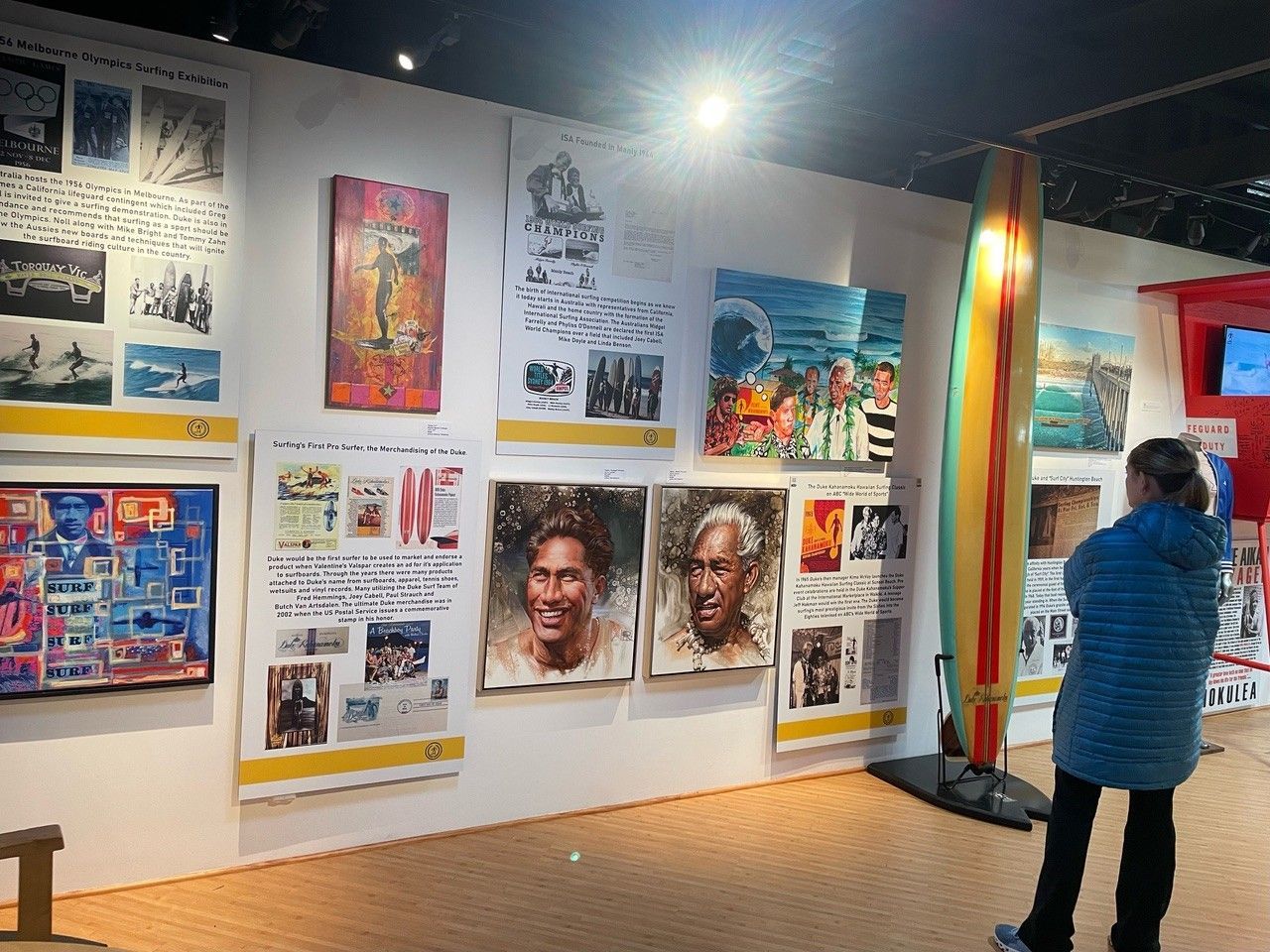 A person viewing an art exhibit featuring posters, paintings, and a colorful surfboard.