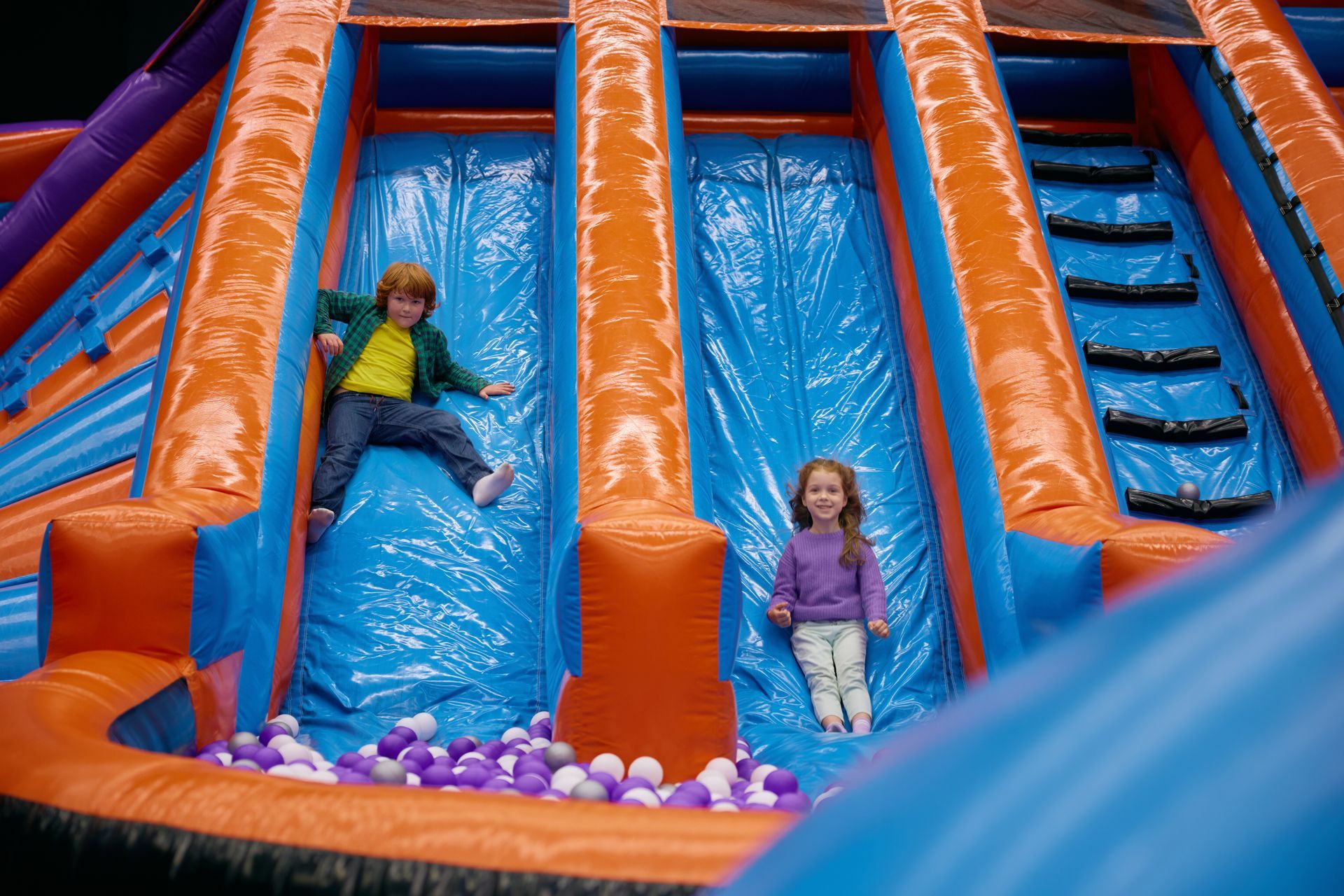 Children on a blue inflatable slide with orange rails and purple balls at the bottom