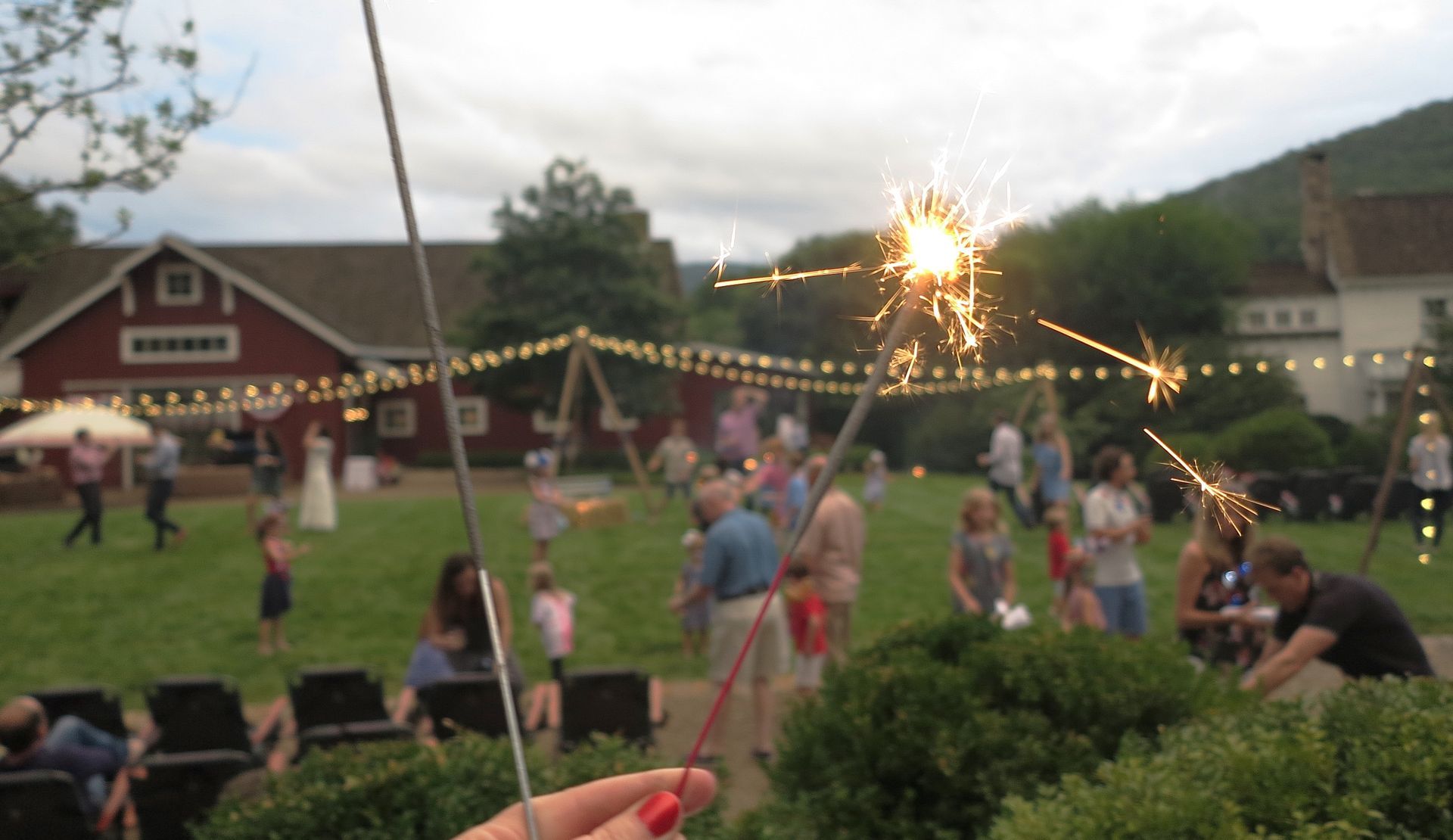 People gathered on a grassy lawn at dusk, watching a glowing sparkler on a string with red barns in back.