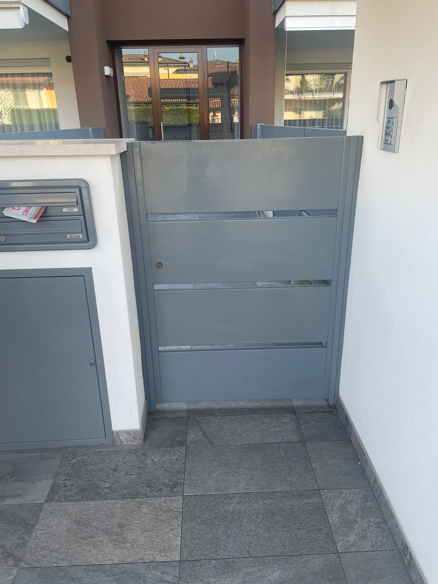 Cancello grigio e cortile piastrellato all'ingresso di un'abitazione, con cassetta delle lettere sulla sinistra e muri bianchi.