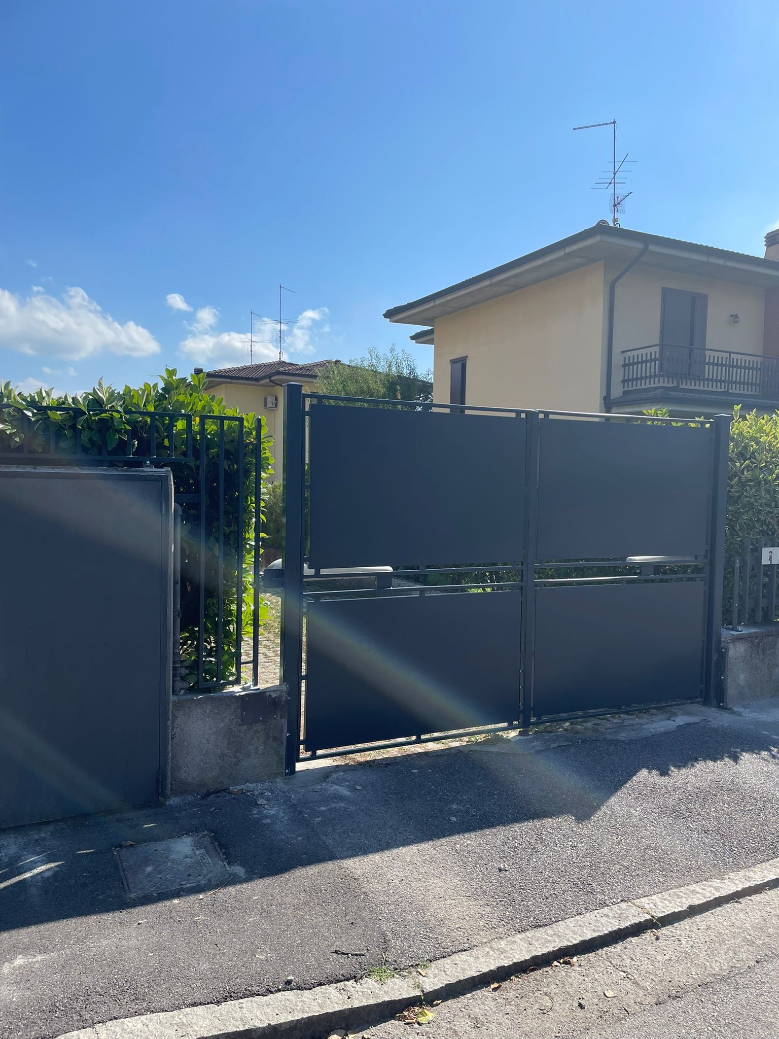 Vialetto di accesso residenziale con cancello e recinzione neri accanto a una casa a due piani color crema sotto un cielo azzurro