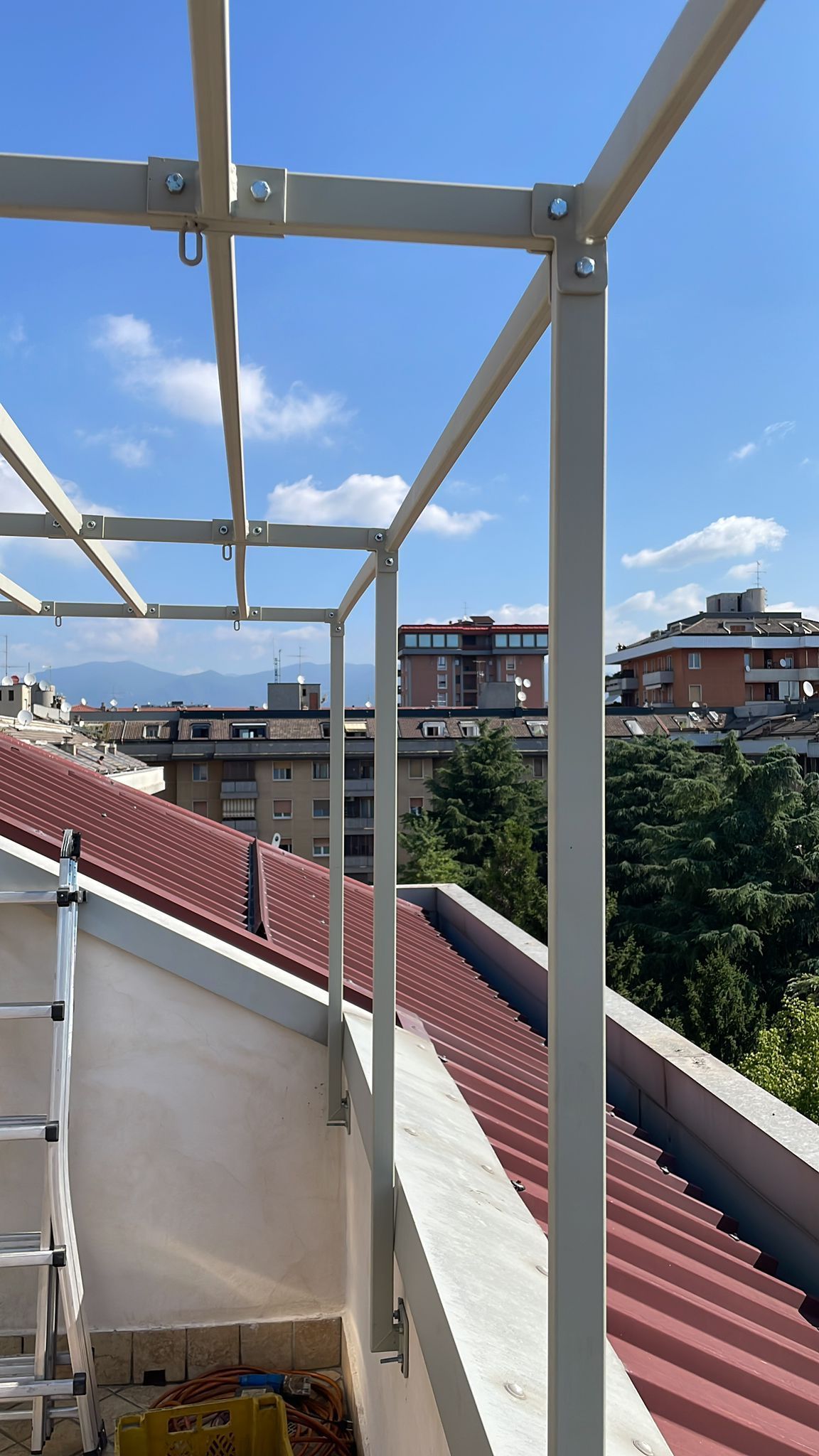 Terrazza sul tetto con struttura in metallo bianco, tetto rosso e palazzi cittadini sotto un cielo blu con nuvole.