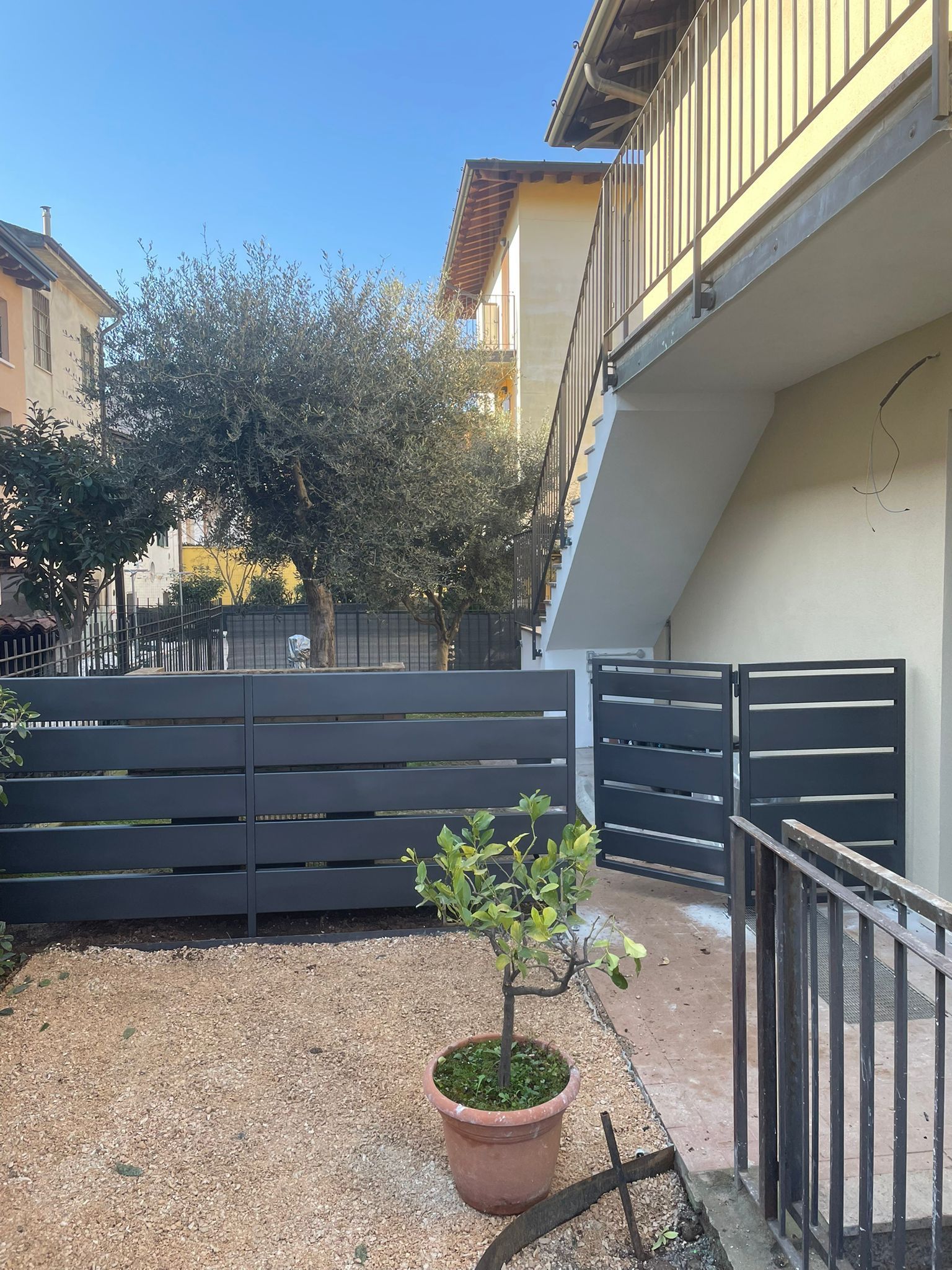 Cortile con recinzione grigia, pianta in vaso, pavimento in ghiaia e una scala accanto a una casa