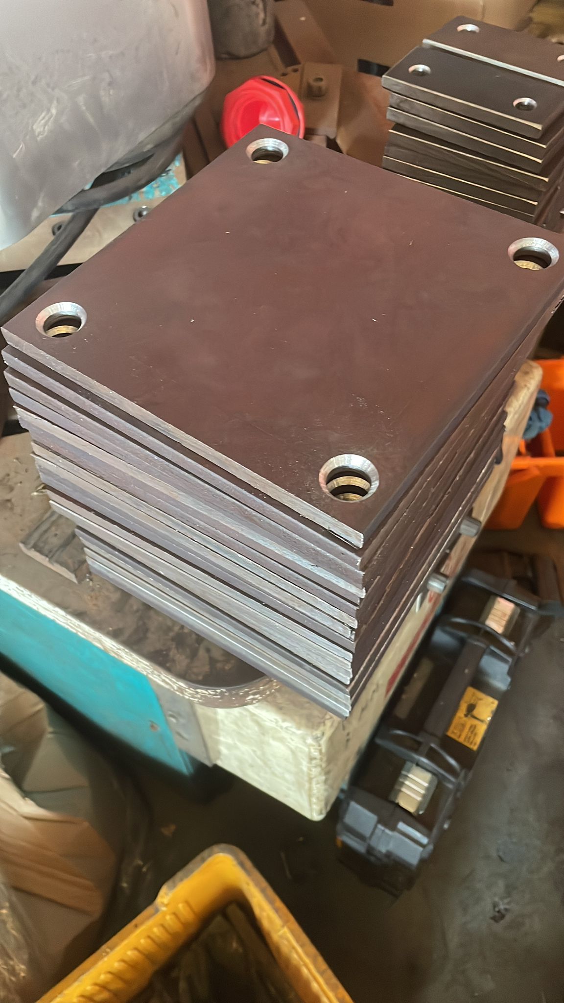 Pila di piastre metalliche marroni e grigie con fori agli angoli su un banco da lavoro industriale.