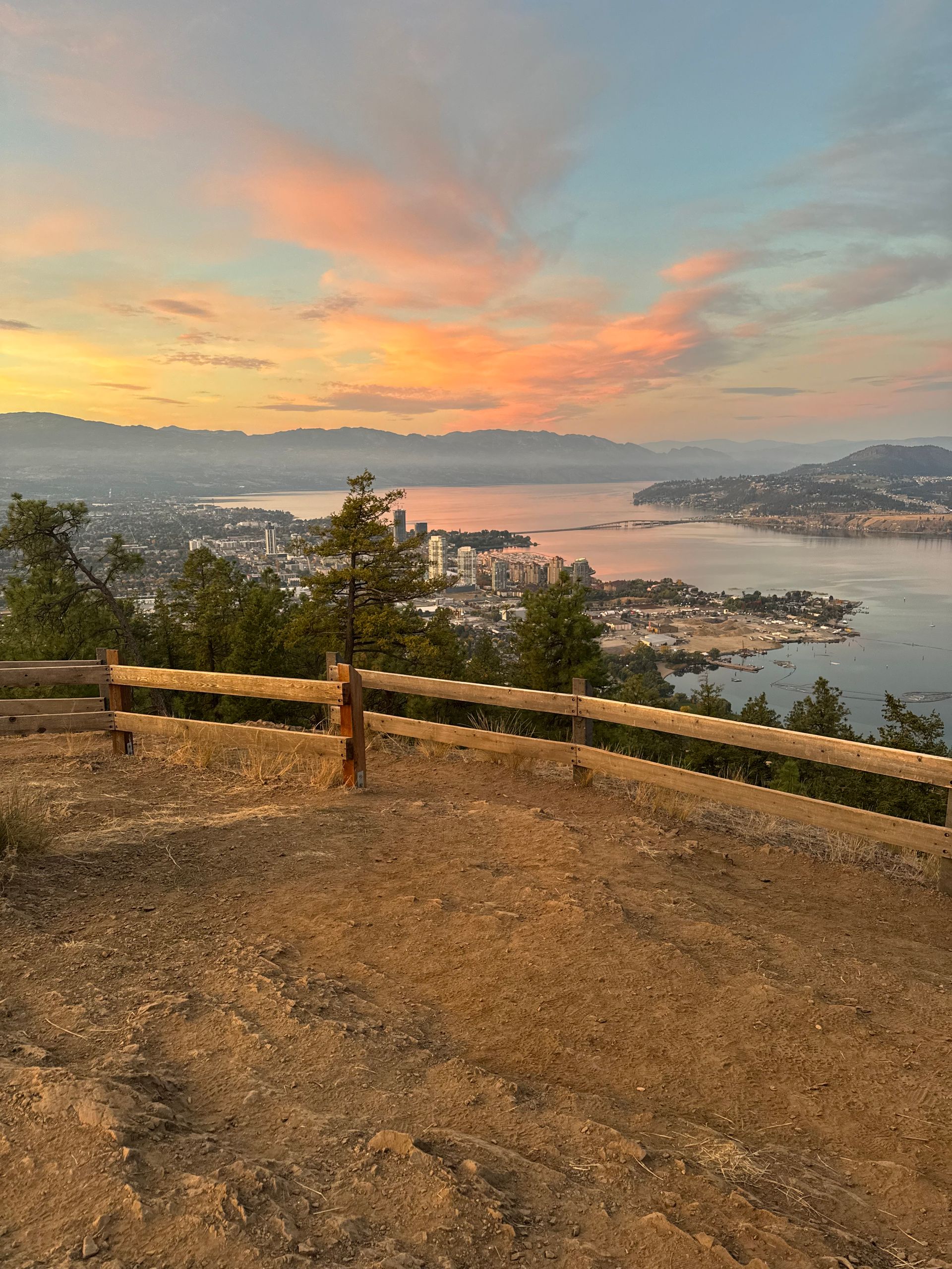 Sunset over a city and lake, viewed from a hilltop with a wooden fence. Pink and orange sky.
