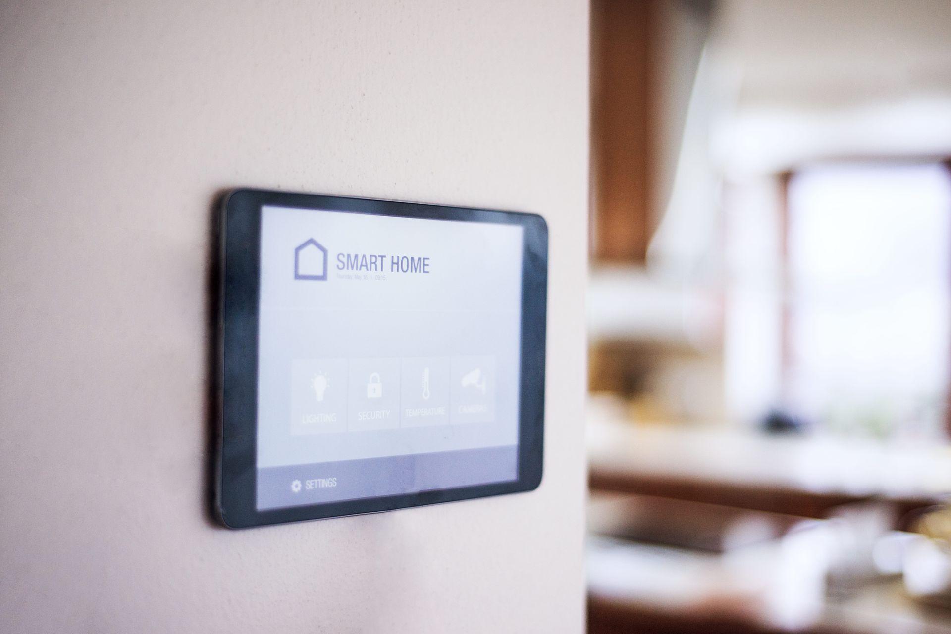 Smart home control panel mounted on a beige wall, displaying icons and text in a bright kitchen.