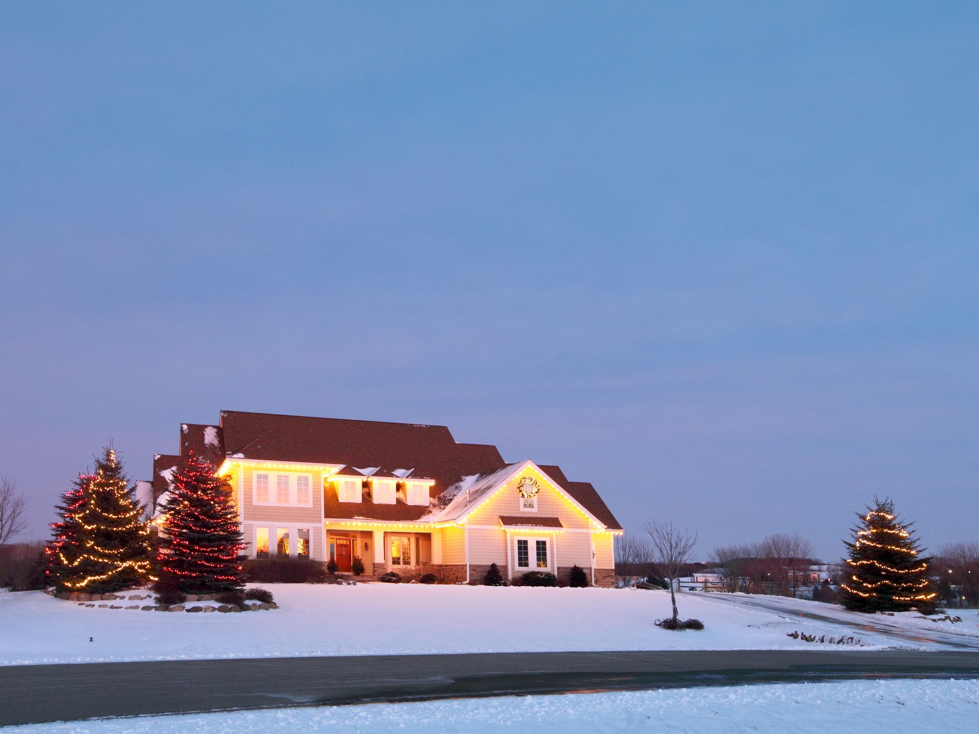 Snowy house at dusk, decorated with Christmas lights on trees and roof.