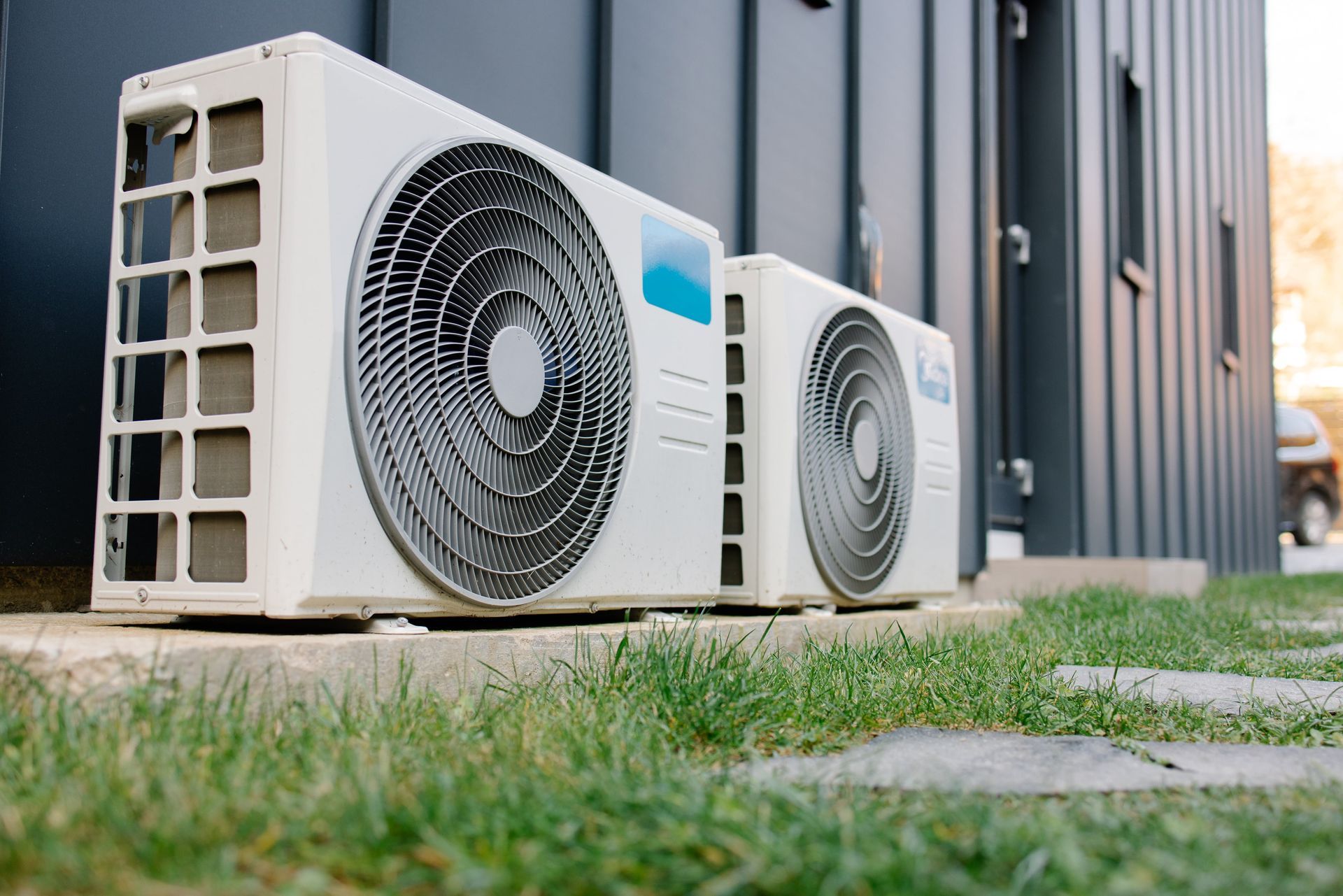 Two white air conditioning units outside, on a concrete slab near a building with gray siding, and green grass.