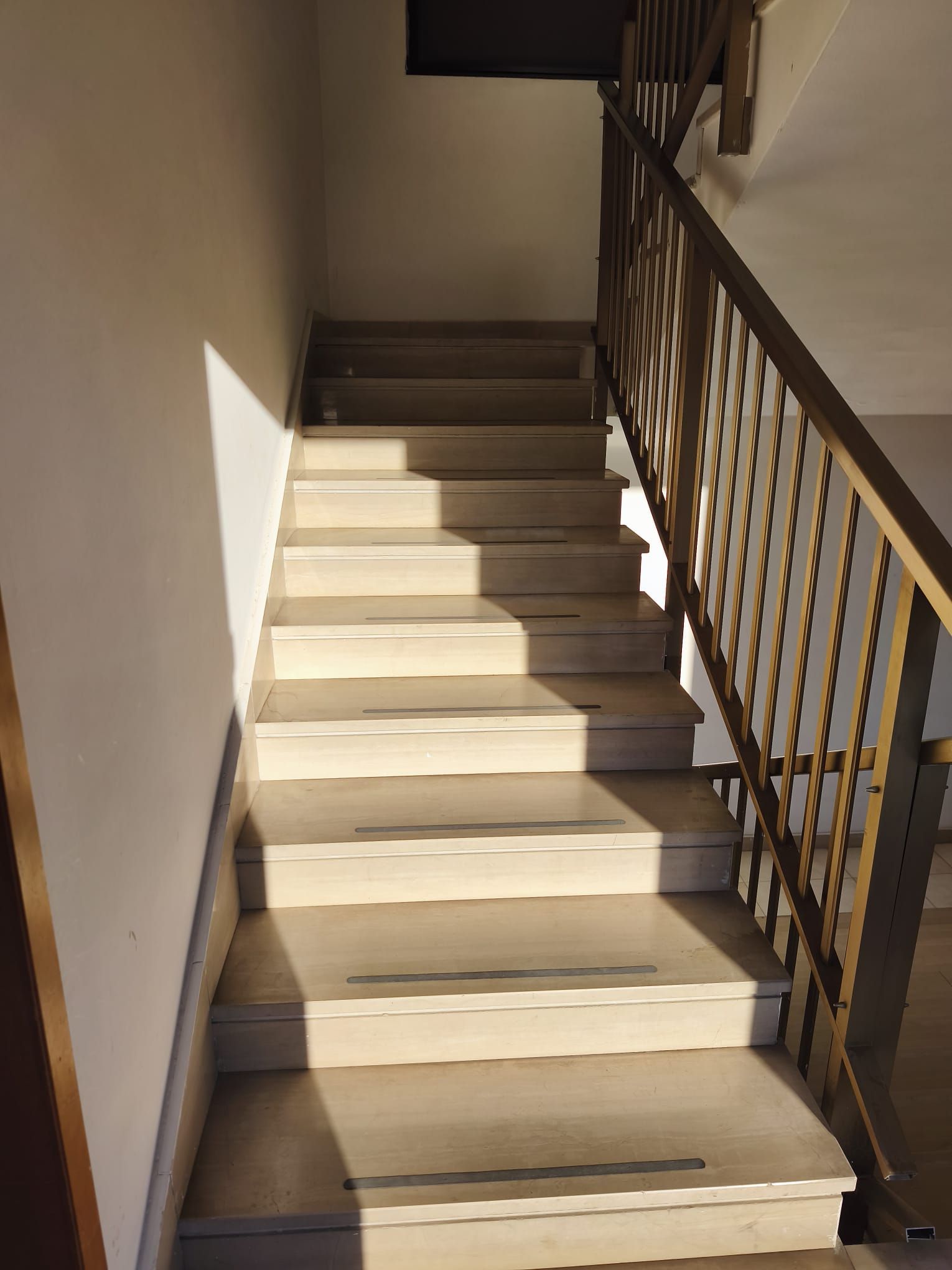 Scala interna con gradini beige e ringhiera in metallo marrone, illuminata dalla luce del sole.