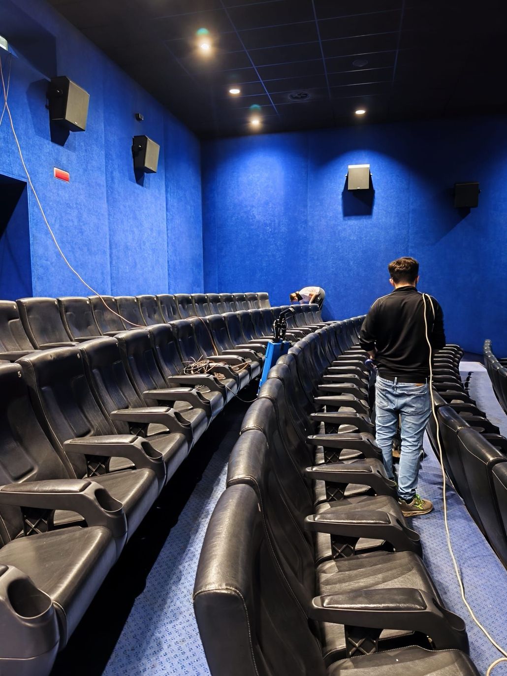 Una sala cinematografica con pareti blu, file di poltrone nere e una persona che cammina lungo il corridoio.