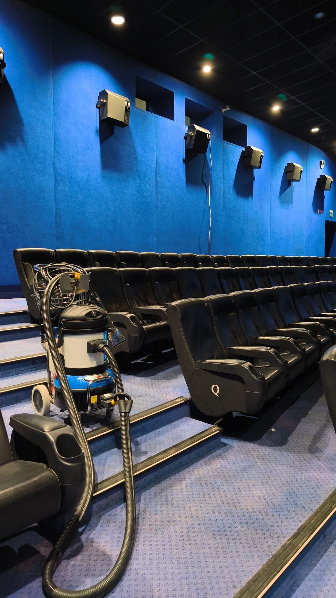 Una sala cinematografica vuota con pareti blu, file di poltrone nere e un treppiede per la macchina fotografica nel corridoio.