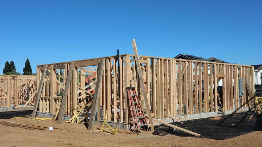 Wood-framed house under construction on a dirt lot, with ladders and building materials visible.