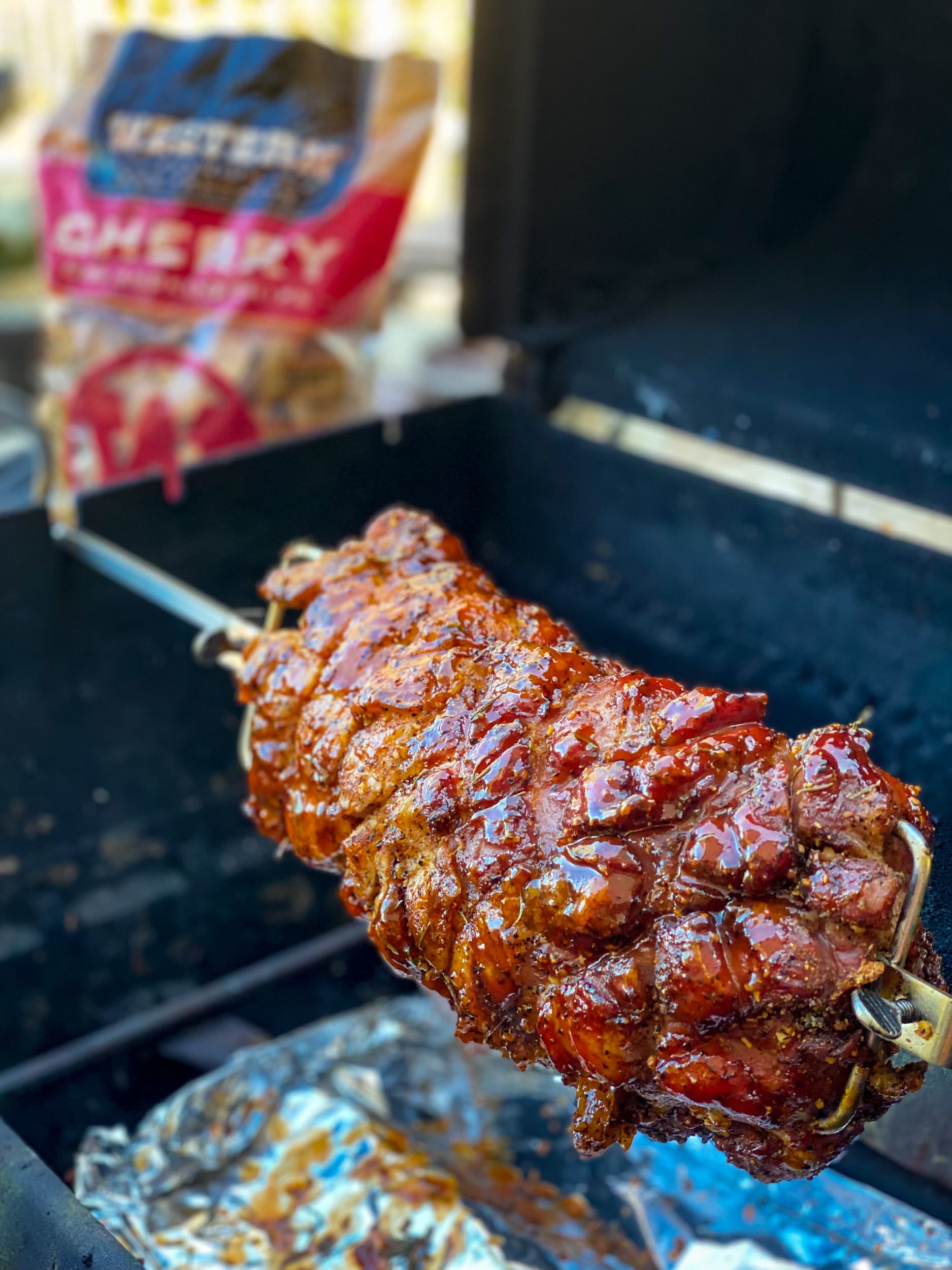 Pork loin cooking on rotisserie grill with cherry chips bag in background