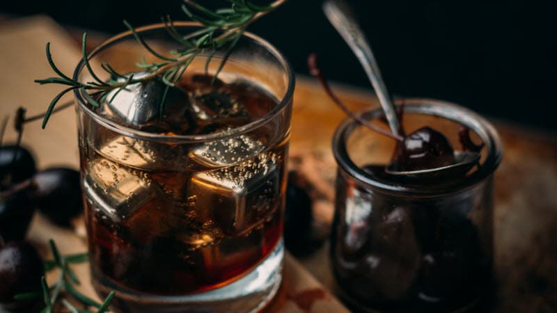 Cocktail with ice and rosemary sprig next to jar of candied cherries