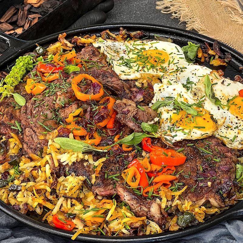 grilled steaks and fried eggs on a bed of hashbrowns on cast iron pan