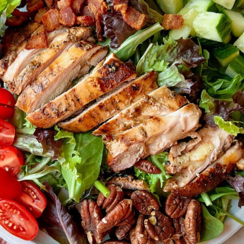 Sliced grilled chicken thighs on bed of lettuce with tomatoes, cucumbers and walnuts