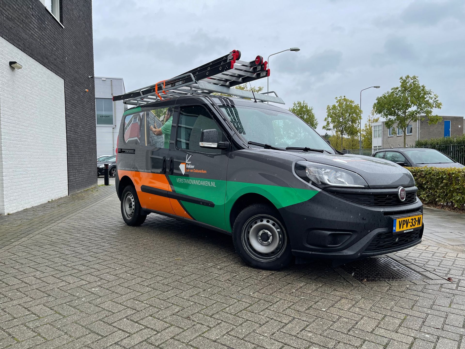 Een busje met een ladder op het dak staat geparkeerd voor een gebouw.