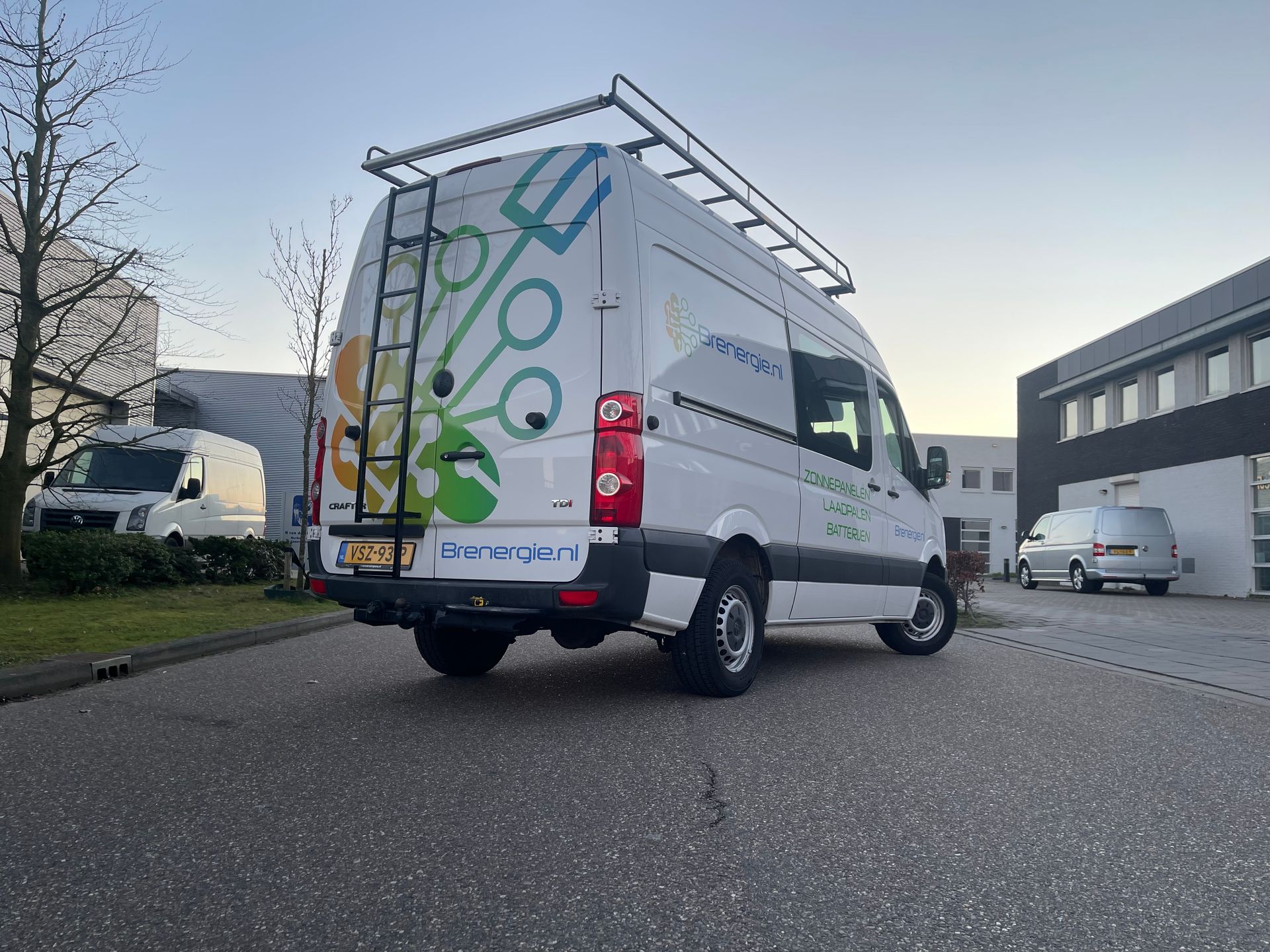 Een wit busje met een ladder achterop staat geparkeerd voor een gebouw.